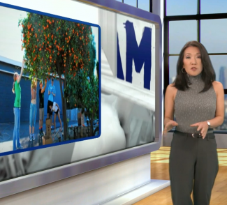 Newscaster stands in front of a screen showing scenes from a harvest